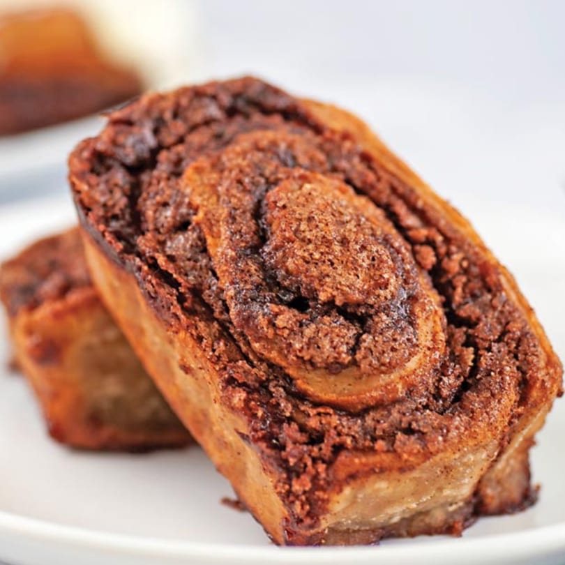 Two golden-brown Crumble & Whisk cinnamon rolls with dark, swirled cinnamon filling on a white plate.