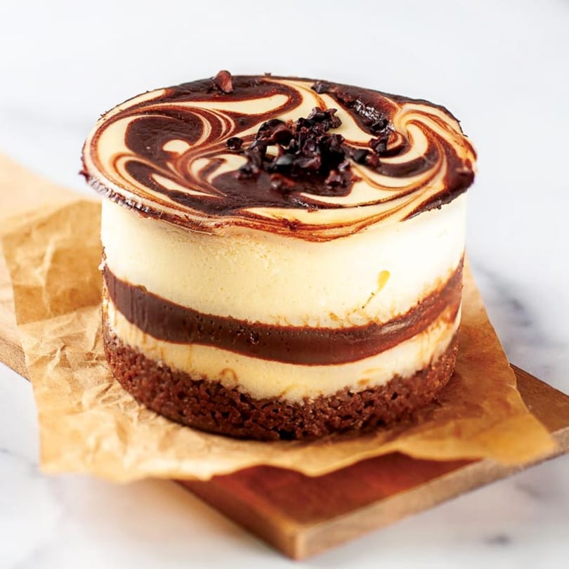A Crumble & Whisk cheesecake with chocolate swirl topping and chocolate chips, on a wooden board.