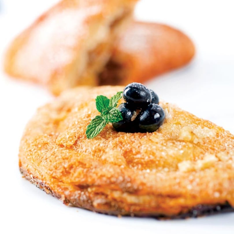 Golden-brown pastry topped with blueberries and a sprig of mint, with another pastry in the background.