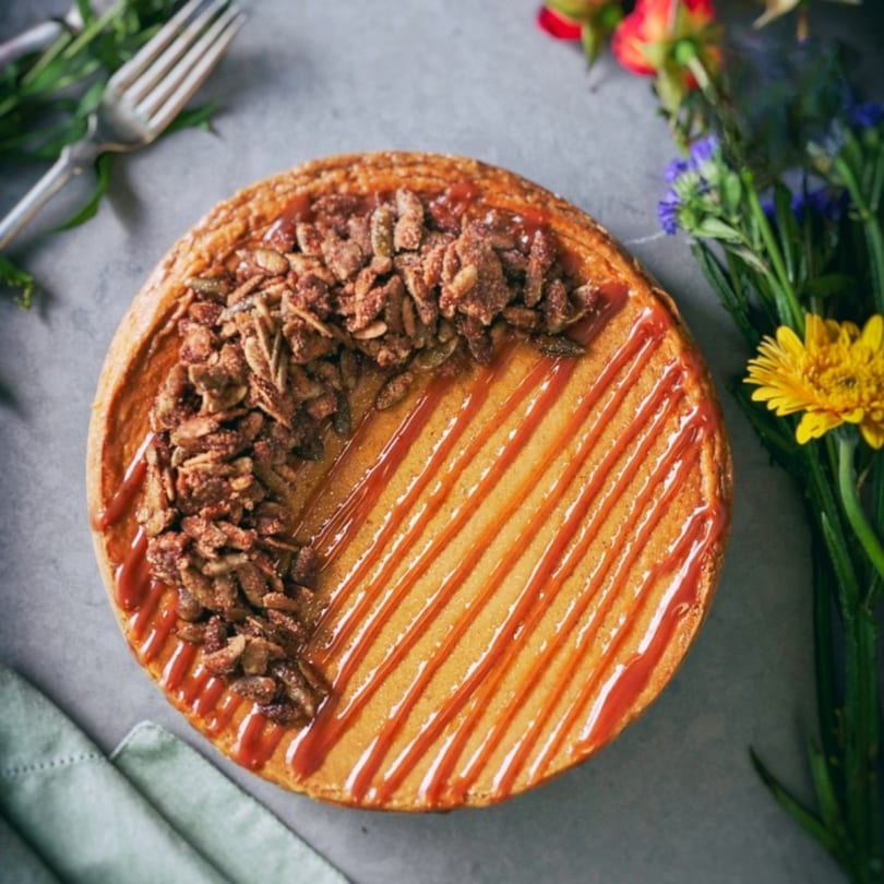 Overhead view of a Crumble & Whisk cheesecake with caramel drizzle and granola, next to flowers.