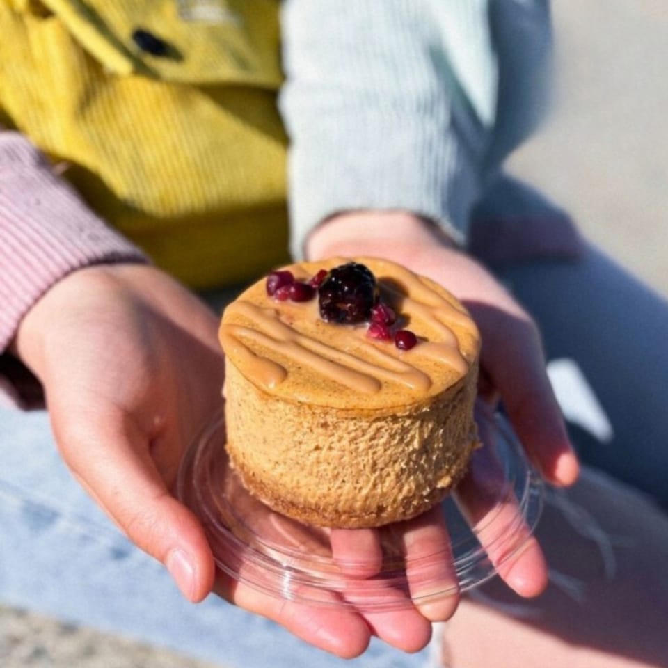 Hands holding a small Crumble & Whisk cheesecake topped with berries and caramel drizzle.