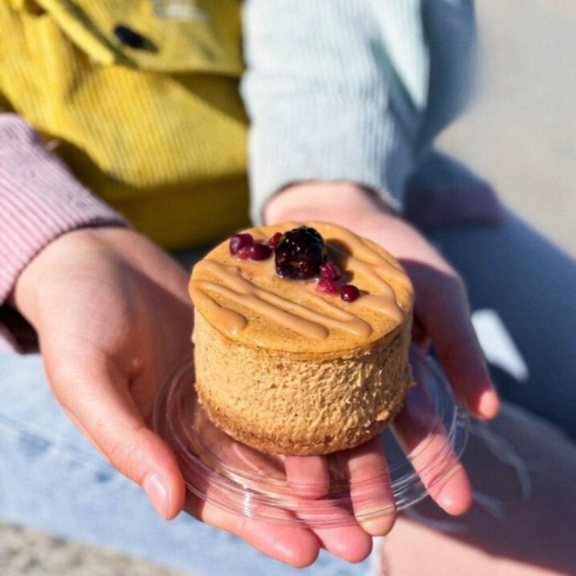 Hands holding a small Crumble & Whisk cheesecake topped with berries and caramel drizzle.